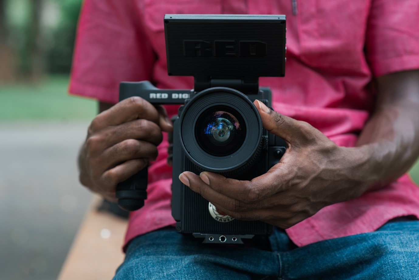 A person wearing a pink shirt and jeans is holding a large professional camera with both hands. The camera's lens and viewfinder are prominent, and the focus is on the camera's details.