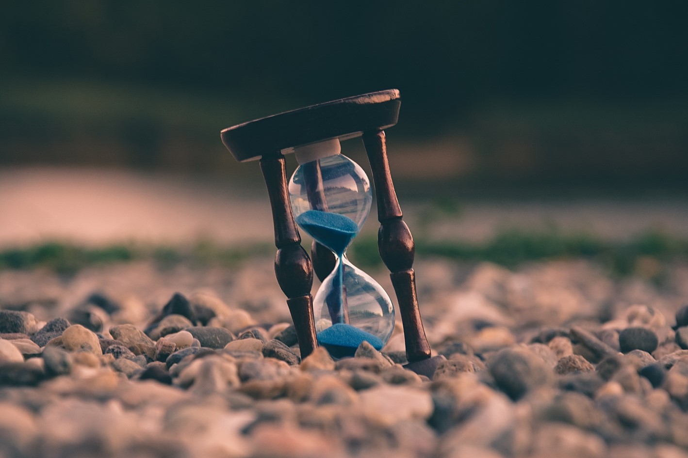 An hourglass with blue sand is resting on a bed of small, smooth stones. The hourglass is slightly tilted, and the background is blurred with earthy tones.