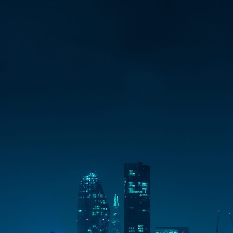 A cityscape at night with skyscrapers illuminated in blue tones, reflecting on the river below. Boats are docked along the water, and festive lights line the trees on the riverbank.